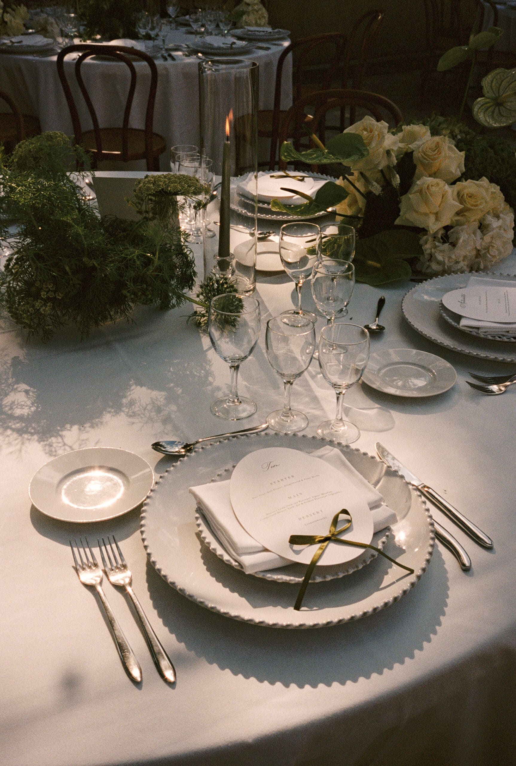 Table de dîner élégante : vaisselle perlée, couverts argent, roses blanches et verdure. Le menu oval "Tom" est lié par un ruban vert.