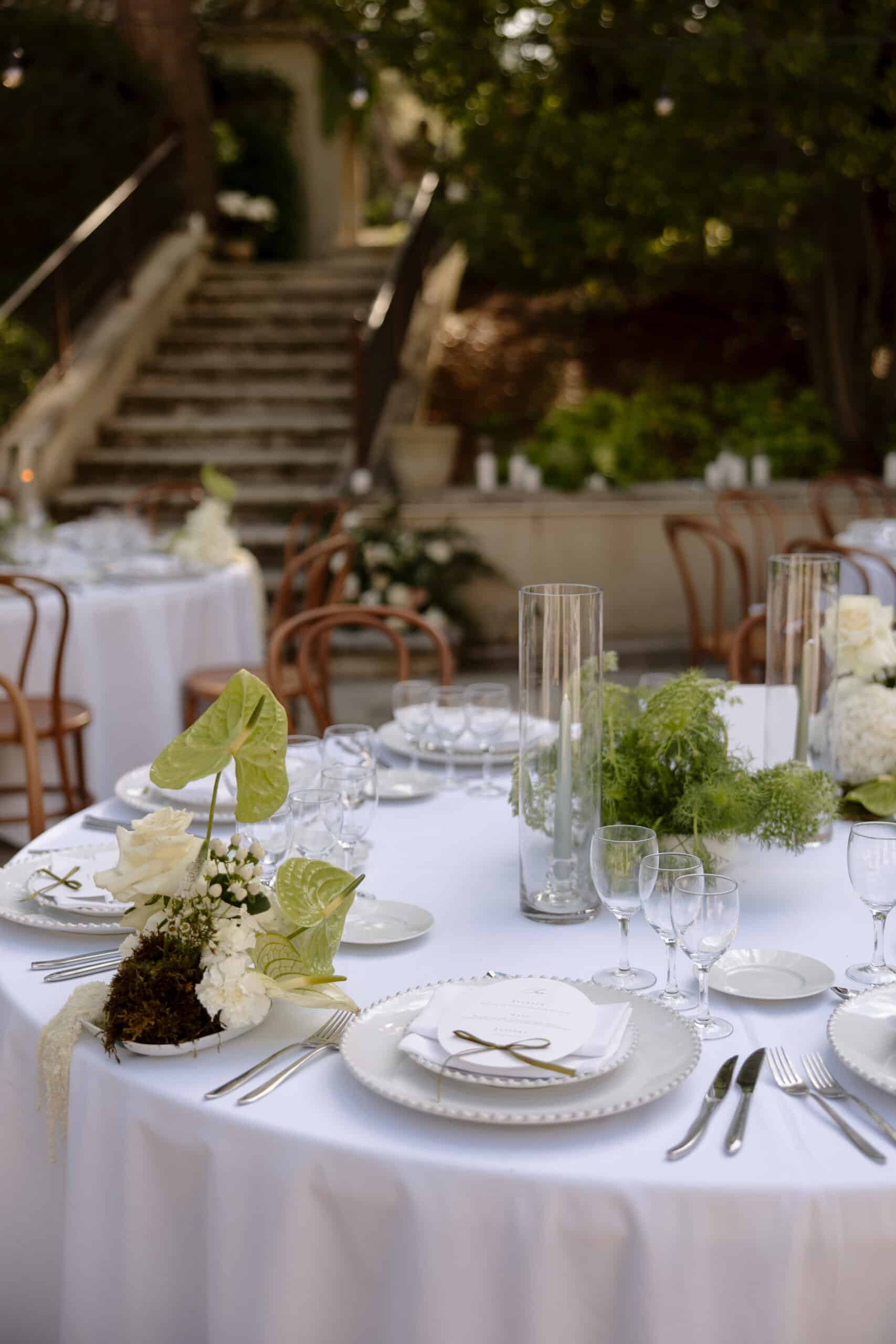 Table de réception de mariage dressée en extérieur. Décoration florale vert et blanc avec roses, Anthurium et mousses. Escaliers de pierre en arrière-plan.