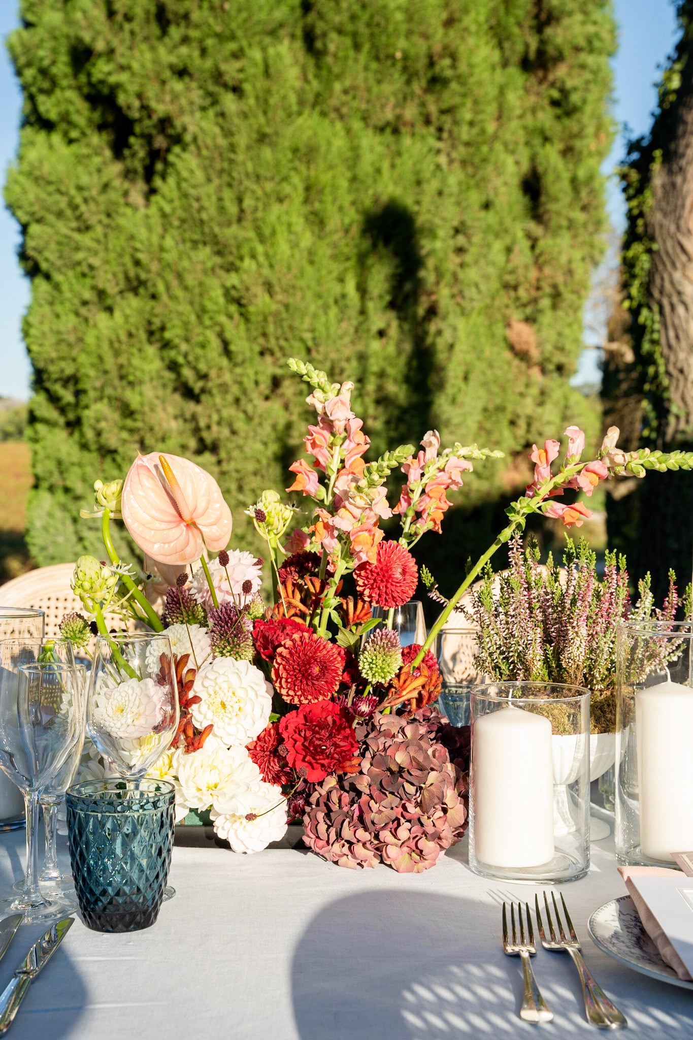 Somptueux centre de table d'automne en extérieur : anthurium rose, dahlias rouges et blancs, hortensias bordeaux, gueules de loup. Couverts et bougies.